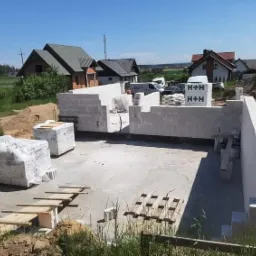 Budowa domu z betonu komórkowego: wzniesione ściany fundamentowe z widocznymi paletami i zapasem materiałów budowlanych H+H w tle, osiedle domów jednorodzinnych.