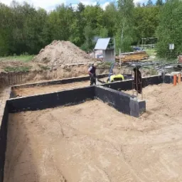 Przygotowany wykop i zbrojone, zaizolowane fundamenty domu jednorodzinnego, widoczny pracownik budowlany, stosy piasku i materiałów budowlanych.