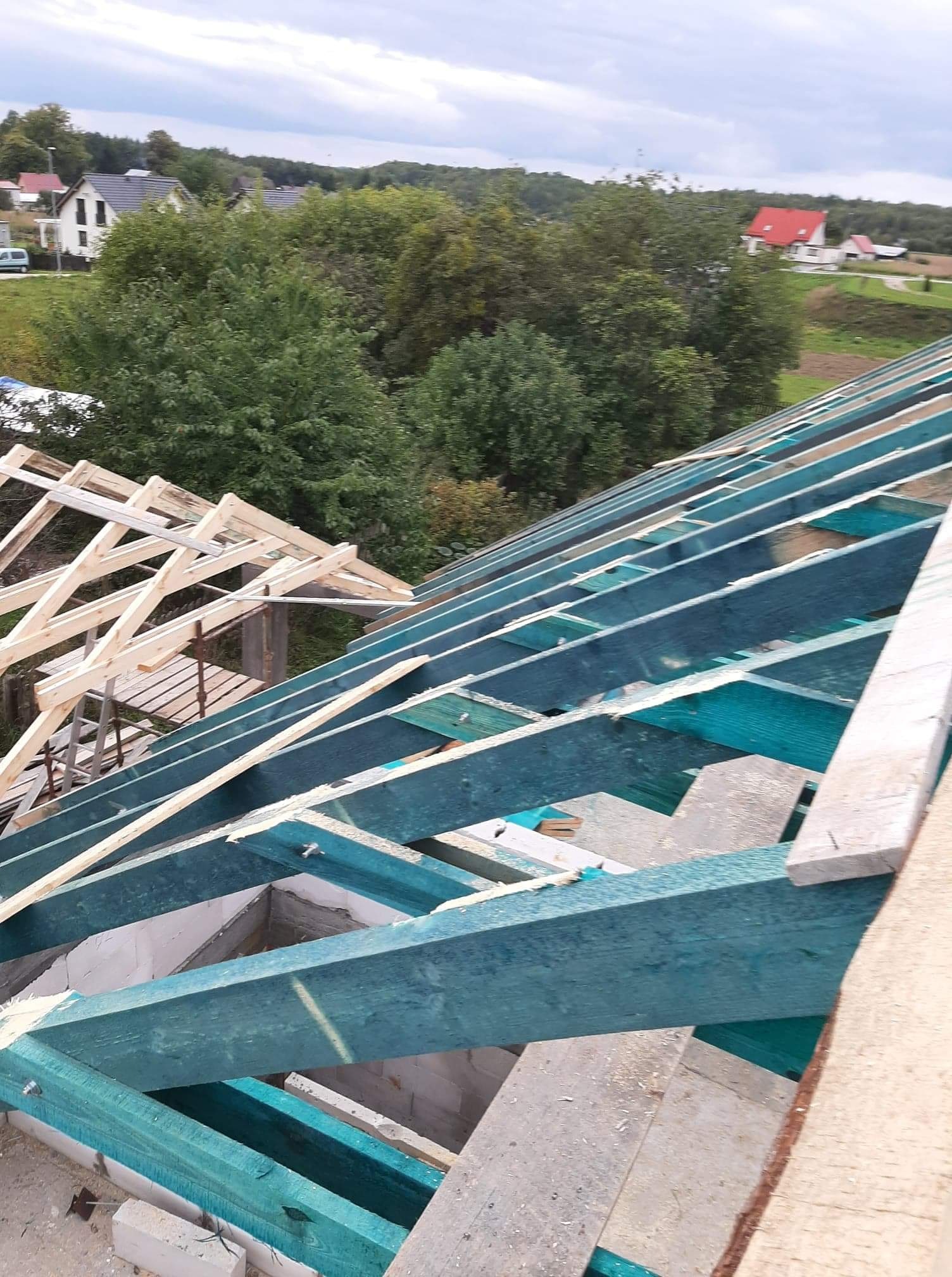 Konstrukcja drewniana dachu w trakcie budowy, widoczne impregnowane elementy w kolorze turkusowym, w tle zabudowa jednorodzinna i zieleń.