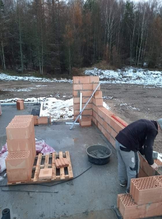 Wznoszenie ściany z bloczków ceramicznych Porotherm na fundamencie w zimowej scenerii, widoczna zaprawa w wiadrze i paleta z bloczkami.