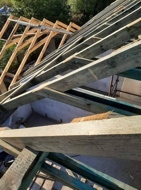 Konstrukcja drewniana dachu w trakcie budowy, widoczne elementy więźby dachowej i fragment murowanej ściany w tle.