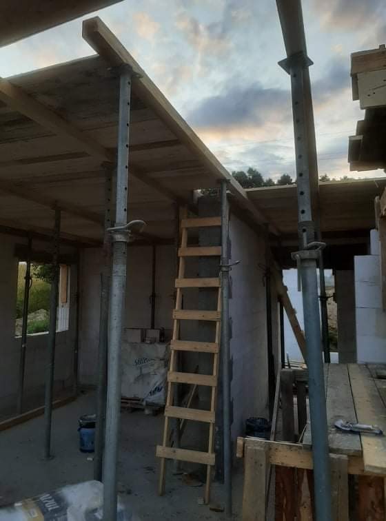 Wnętrze w trakcie budowy domu z widocznym szalunkiem stropu podpartym stalowymi stemplami, drewniana drabina oparta o ścianę z bloczków.
