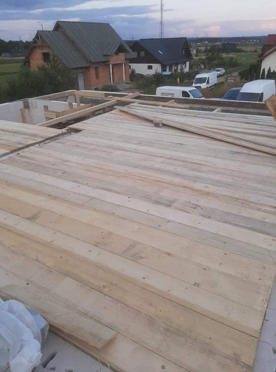 Wiązanie drewnianych elementów konstrukcyjnych na wznoszonym budynku, widoczne białe bloczki fundamentowe i zabudowa w tle.