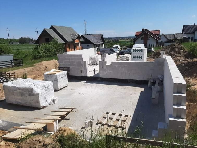 Budowa domu z betonu komórkowego: wzniesione ściany fundamentowe z widocznymi paletami i zapasem materiałów budowlanych H+H w tle, osiedle domów jednorodzinnych.