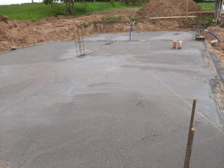 Wylany fundament z wystającymi prętami zbrojeniowymi, widoczne niebieskie rury instalacyjne i wytyczone sznurkiem linie, grunt wokół wykopany.