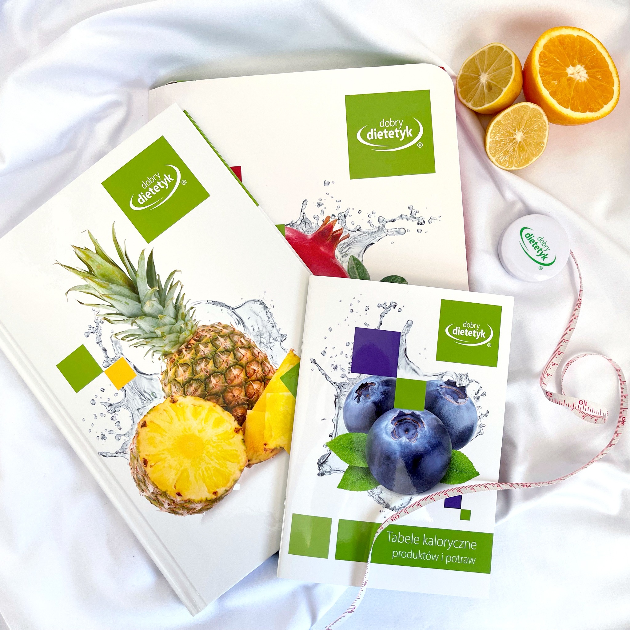 Trzy książki z owocami i logo na okładkach leżą na białej, delikatnie pofałdowanej tkaninie. Obok leżą połówki cytryny i pomarańczy oraz miarka krawiecka z logo.