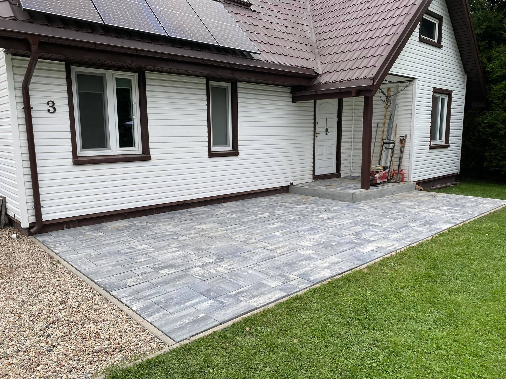 Nowoczesny podjazd z szarej kostki brukowej ułożonej przed domem z białym sidingiem i brązowym dachem. Widoczne panele słoneczne na dachu, numer domu '3' oraz narzędzia opierające się o ścianę...
