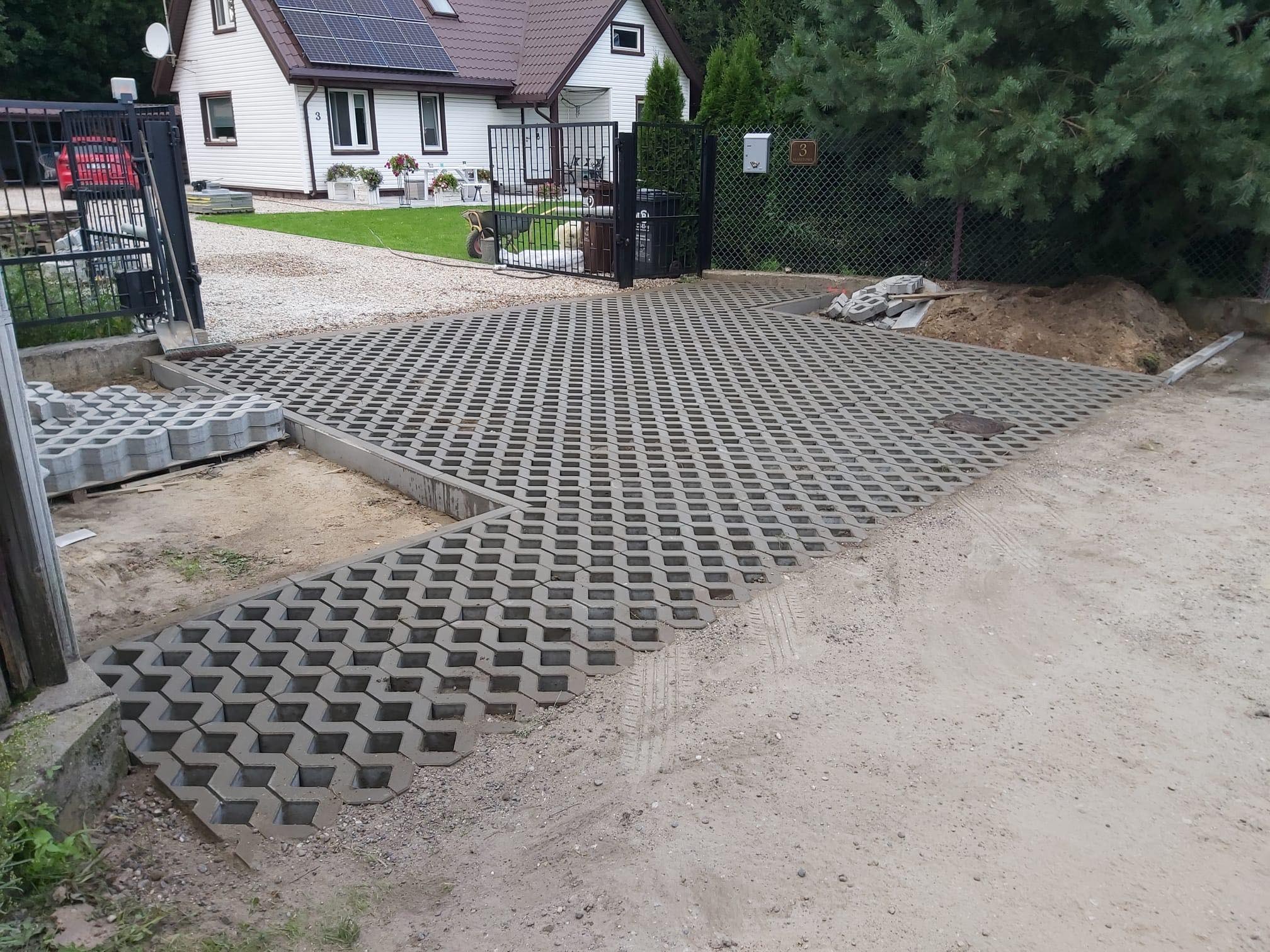 Fragment podjazdu z częściowo ułożoną ażurową kostką betonową na piaskowym podłożu, widoczne przygotowane elementy kostki i krawężniki, w tle dom jednorodzinny z panelami słonecznymi.