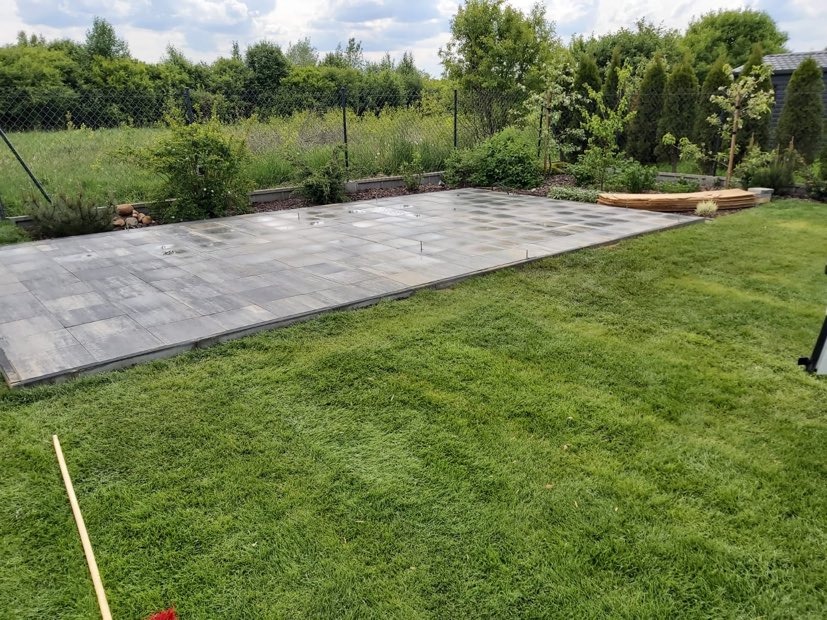 Układanie szarej kostki brukowej na przygotowanym podłożu w ogrodzie, widoczne pręty stabilizujące i paleta z kostką w tle.