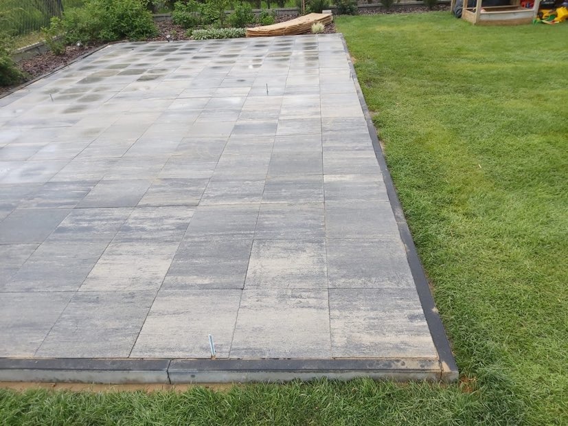 Ułożona nawierzchnia z szarej kostki brukowej, widoczne metalowe szpilki stabilizujące, obrzeża z ciemnej kostki, trawnik i fragment placu zabaw w tle.