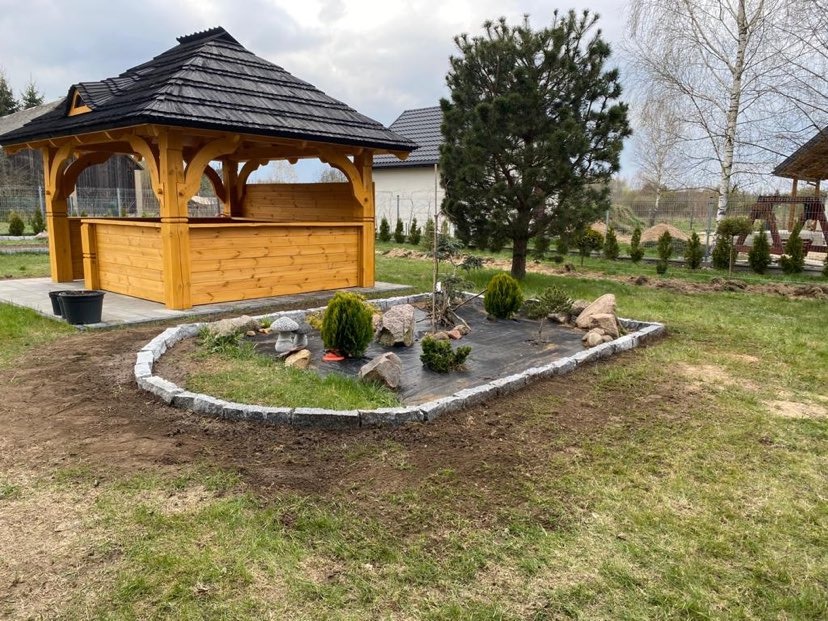 Drewniana altana z czarnym dachem i ogródek wykończony obrzeżem z kostki granitowej, widoczne przygotowane podłoże i rozłożona agrowłóknina.