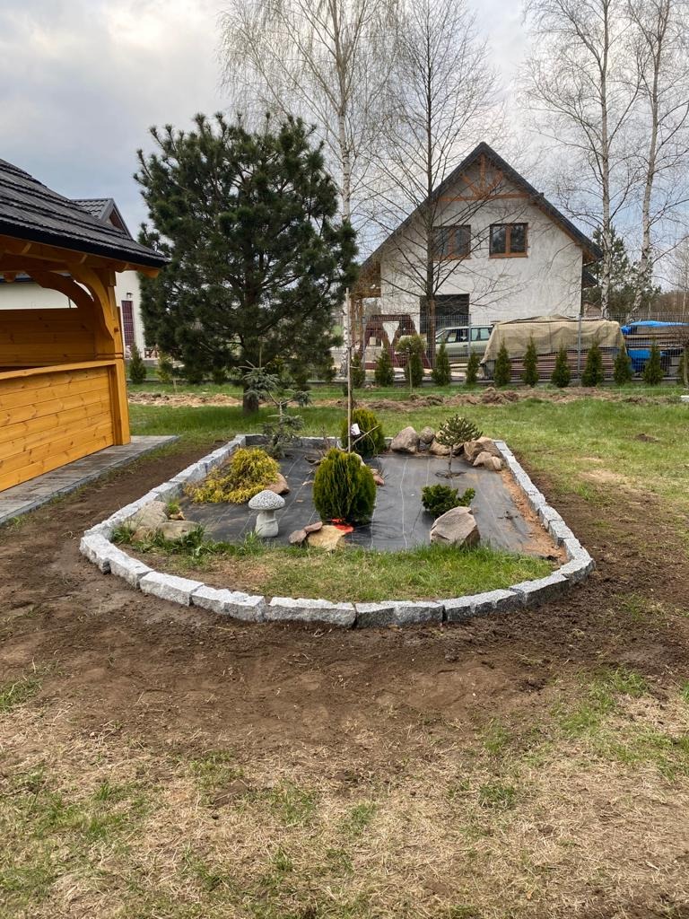 Rabata ogrodowa obłożona kostką granitową, wyłożona agrowłókniną, z nasadzeniami iglaków i kamieniami, na tle domu jednorodzinnego i altany.