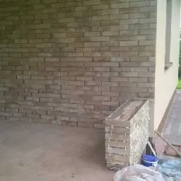 Fragment elewacji budynku z wykończeniem z cegiełek klinkierowych, widoczny stos cegieł, wiadro z farbą, oraz zabezpieczająca folia malarska.