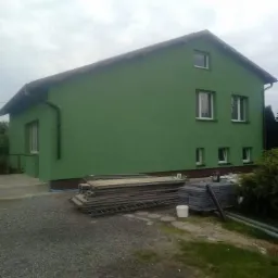 Dom jednorodzinny z zieloną elewacją po malowaniu, widoczne rusztowania i materiały budowlane na posesji, pochmurne niebo.