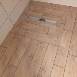 Ułożona podłoga prysznicowa z płytek imitujących drewno, z widocznym odpływem liniowym i krzyżykami dystansowymi w fugach.