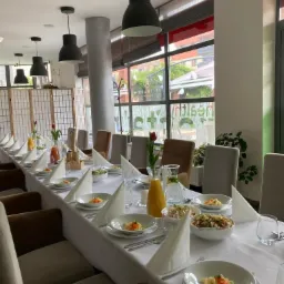 Długi stół nakryty białym obrusem, przygotowany do przyjęcia gości. Widoczne talerze z przystawkami, szklanki, sztućce, serwetki ułożone w trójkąty, dzbanki z sokiem i wodą z cytryną. W tle okno...