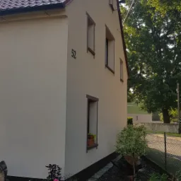 Jasna elewacja domu jednorodzinnego z numerem 52, czerwoną dachówką i brązowymi obramowaniami okien, otoczona zielenią i kwiatami w donicach.