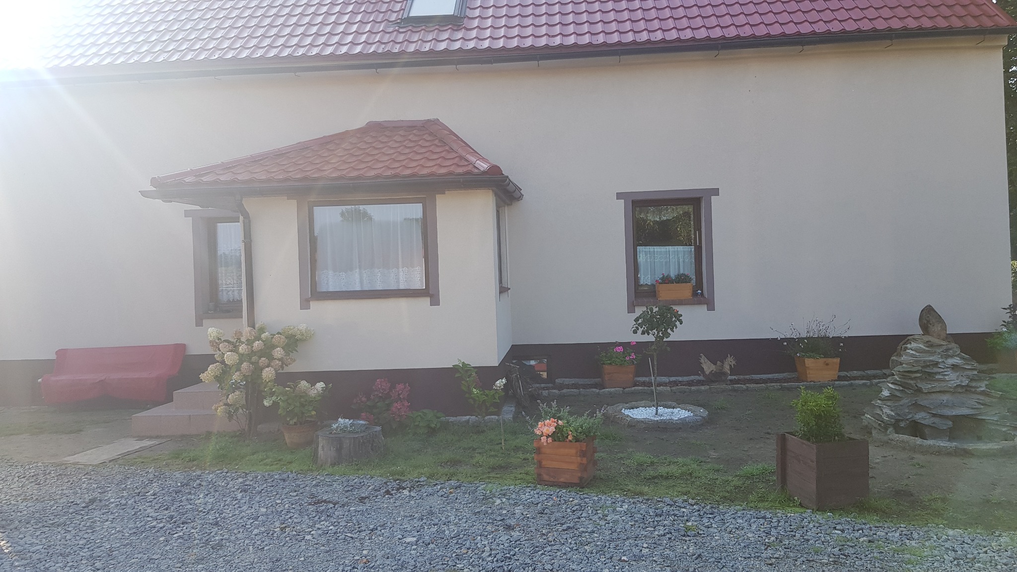 Jasna elewacja domu jednorodzinnego z czerwonym dachem, okna z brązowymi ramami, ozdobne donice z kwiatami i kamienna dekoracja ogrodowa.