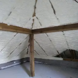 Poddasze z drewnianą konstrukcją krokwiową pokryte grubą warstwą białej pianki izolacyjnej, widoczne niedociągnięcia w aplikacji w górnej części dachu.