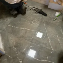 Układanie płytek podłogowych z szarym marmurkowym wzorem, widoczne narzędzia: klucze, pędzel i fragment osoby w roboczych butach.