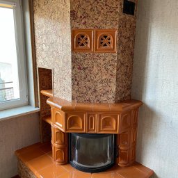 PPHU LEGAM POLSKA EUGENIUSZ LABUDA - Kafelkowy piec w kolorze karmelowym z ozdobnymi kratkami wentylacyjnymi, obudowany korkiem, w rogu pomieszczenia z oknem.