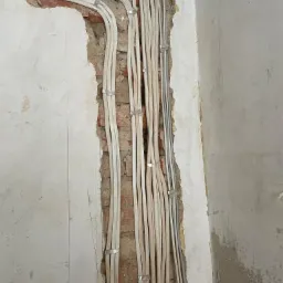 Wiązka przewodów elektrycznych przymocowana metalowymi uchwytami do ściany z fragmentami odsłoniętej cegły podczas prac instalacyjnych.