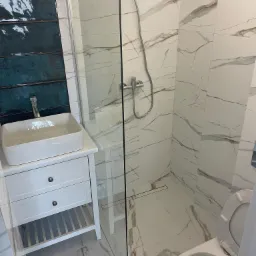 Nowoczesna łazienka po remoncie: białe marmurowe płytki na ścianach i podłodze, kabina prysznicowa ze szklaną ścianką, umywalka nablatowa na białej szafce z szufladami oraz toaleta z otwartą klapą.