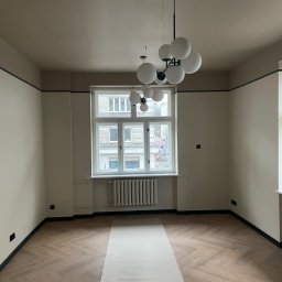 PPHU LEGAM POLSKA EUGENIUSZ LABUDA - Pokój po malowaniu ścian na beżowo z ciemnymi akcentami przy suficie i podłodze, parkiet ułożony w jodełkę zabezpieczony papierem malarskim, nowoczesna lampa wisząca z białymi kulami.