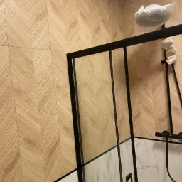 Łazienka z drewnopodobnymi płytkami ułożonymi w jodełkę na ścianie, kabina prysznicowa z czarnymi profilami, armatura zabezpieczona folią ochronną.