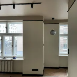 Wnętrze w trakcie malowania: beżowe ściany z czarnymi akcentami, zabezpieczona podłoga w jodełkę, puszka farby na parapecie, widok na miejski krajobraz przez okno.