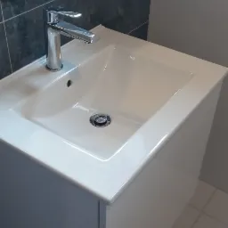 Biała umywalka z szafką w nowoczesnej łazience, chromowana bateria i okrągłe lustro z ciemną obwódką na tle szarych i białych płytek.