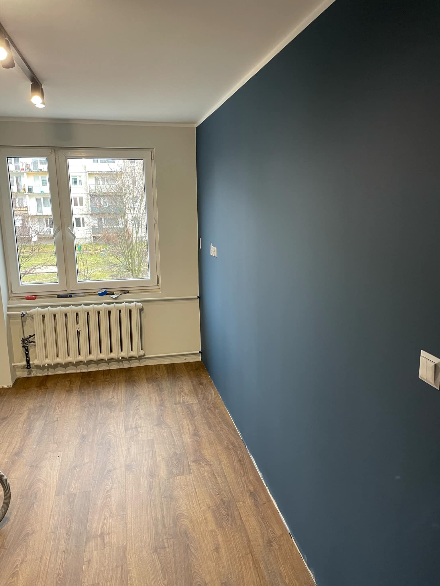 Pokój po malowaniu: ściana w kolorze głębokiego granatu, drewnopodobna podłoga, białe okno z widokiem na blok, biały grzejnik z narzędziami malarskimi na parapecie.