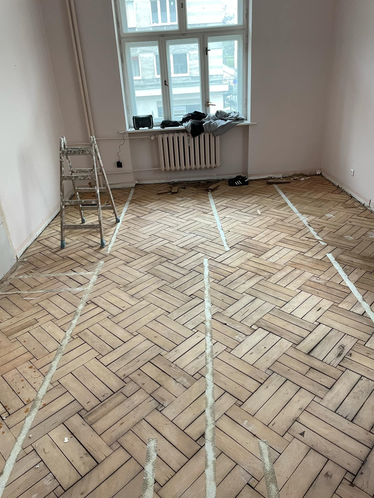 Pokój w trakcie renowacji podłogi parkietowej ułożonej w jodełkę, z widocznymi liniami wyznaczającymi obszary do naprawy lub wymiany, drabina malarska w rogu, okno z grzejnikiem i ubraniami...