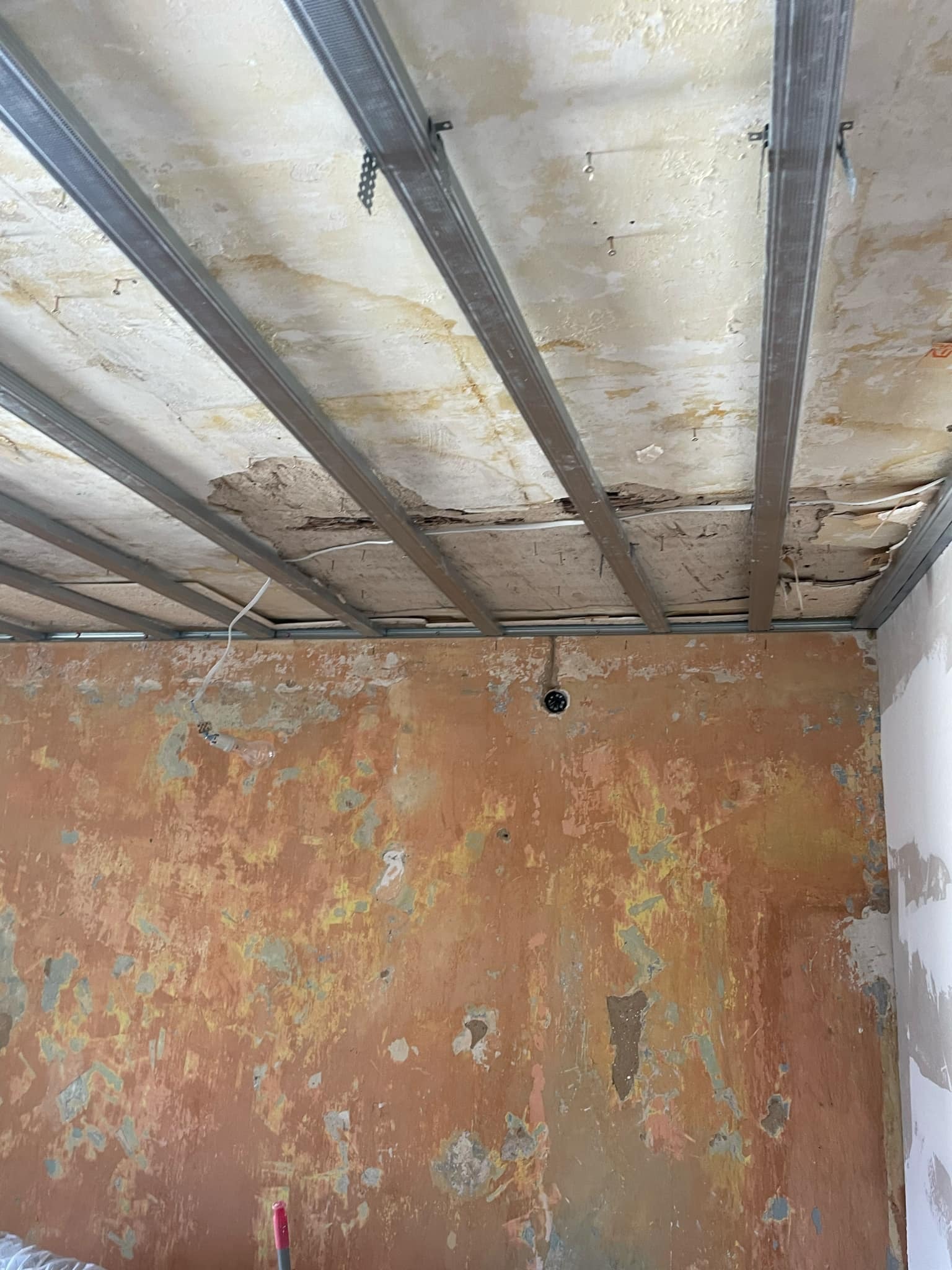 Wnętrze w trakcie remontu: widoczne metalowe profile sufitowe, odsłonięty, zniszczony sufit z widocznymi śladami wilgoci i odpadającą farbą, oraz ściana z resztkami starej farby w odcieniach...