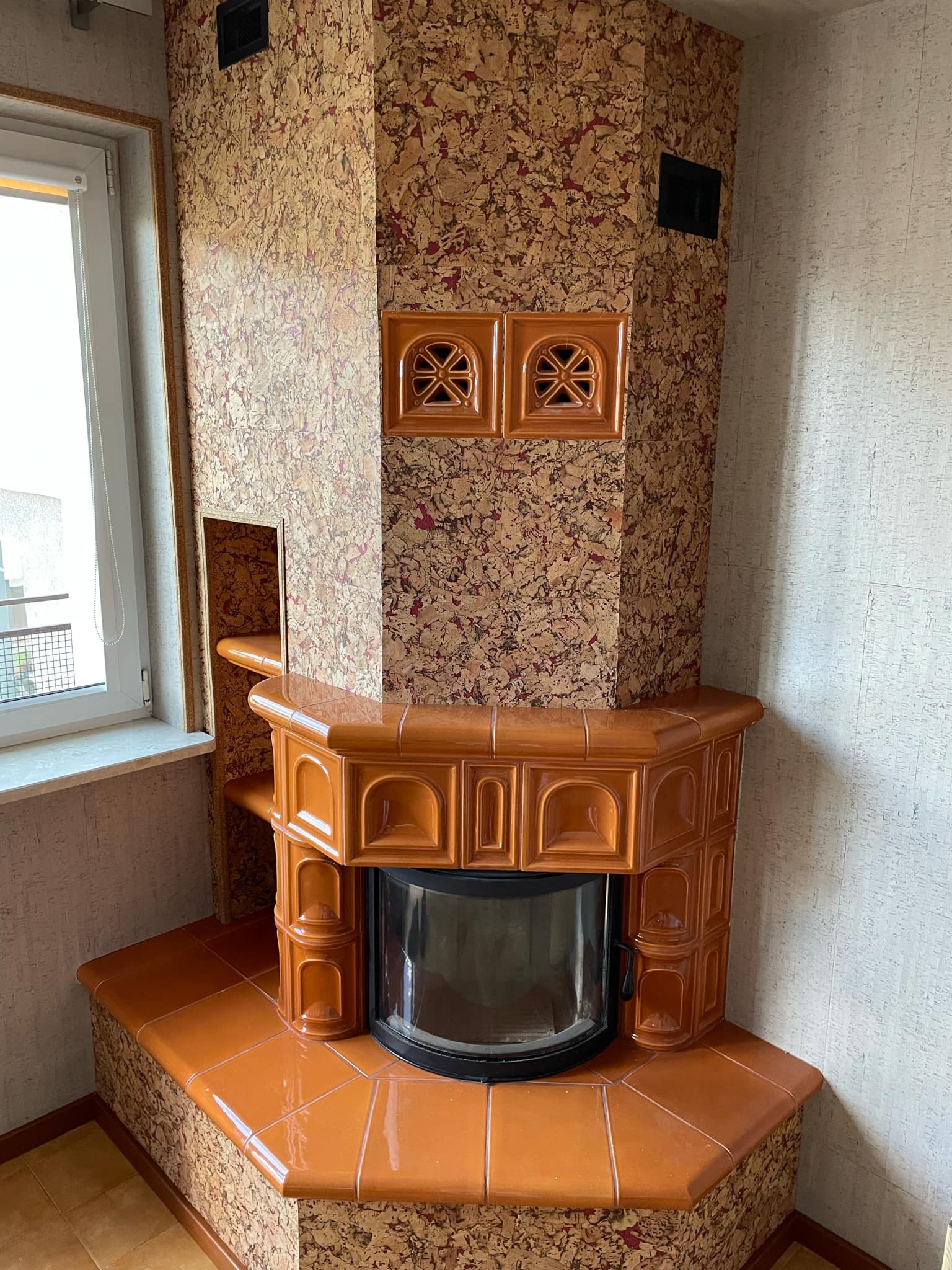 Kafelkowy piec w kolorze karmelowym z ozdobnymi kratkami wentylacyjnymi, obudowany korkiem, w rogu pomieszczenia z oknem.