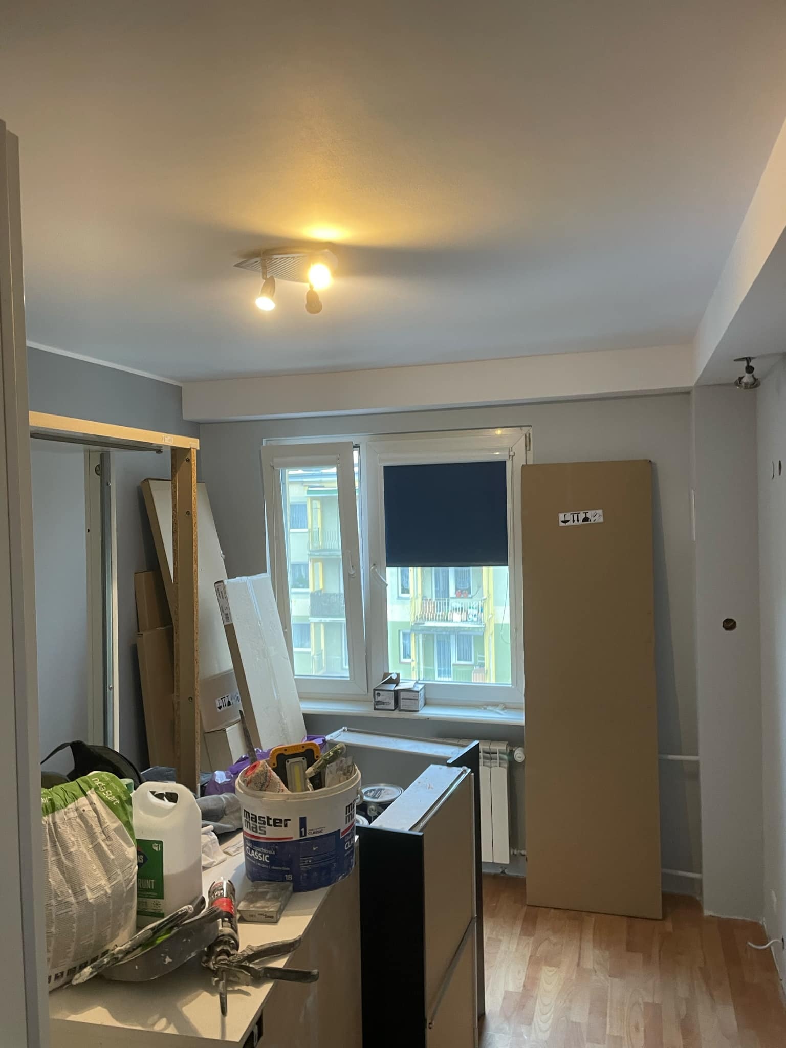 Pokój w trakcie remontu: widoczne narzędzia malarskie, wiadro z farbą Master Mas Classic, kartony, okno z żaluzją i grzejnik.