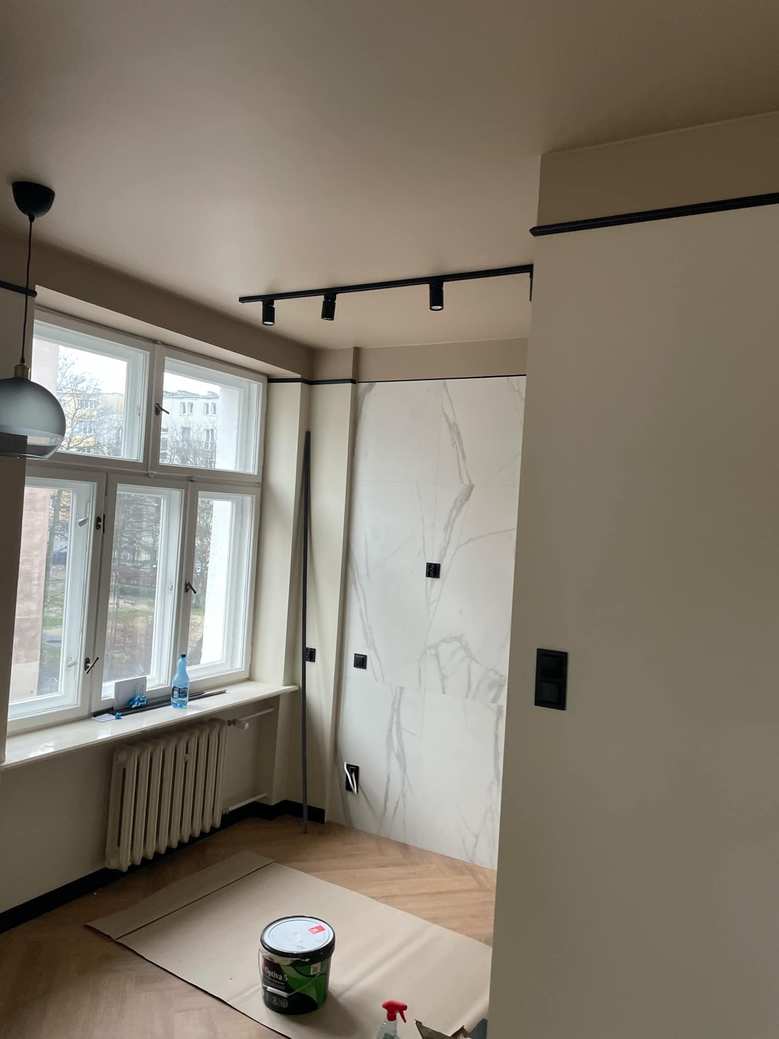 Pokój w trakcie odświeżania: ściany pomalowane na beżowo z czarnymi akcentami, marmurowe płytki na ścianie, zabezpieczona podłoga z parkietem jodełkowym i puszka farby Optiva 5 na kartonie.