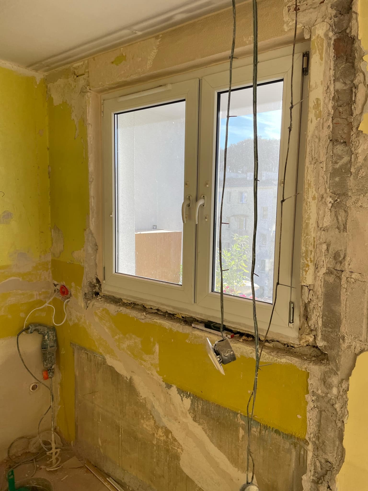 Demontaż okna: Widok na uszkodzone ściany z resztkami żółtej farby, odsłonięty mur, wiszące kable elektryczne i narzędzia, w trakcie prac remontowych.