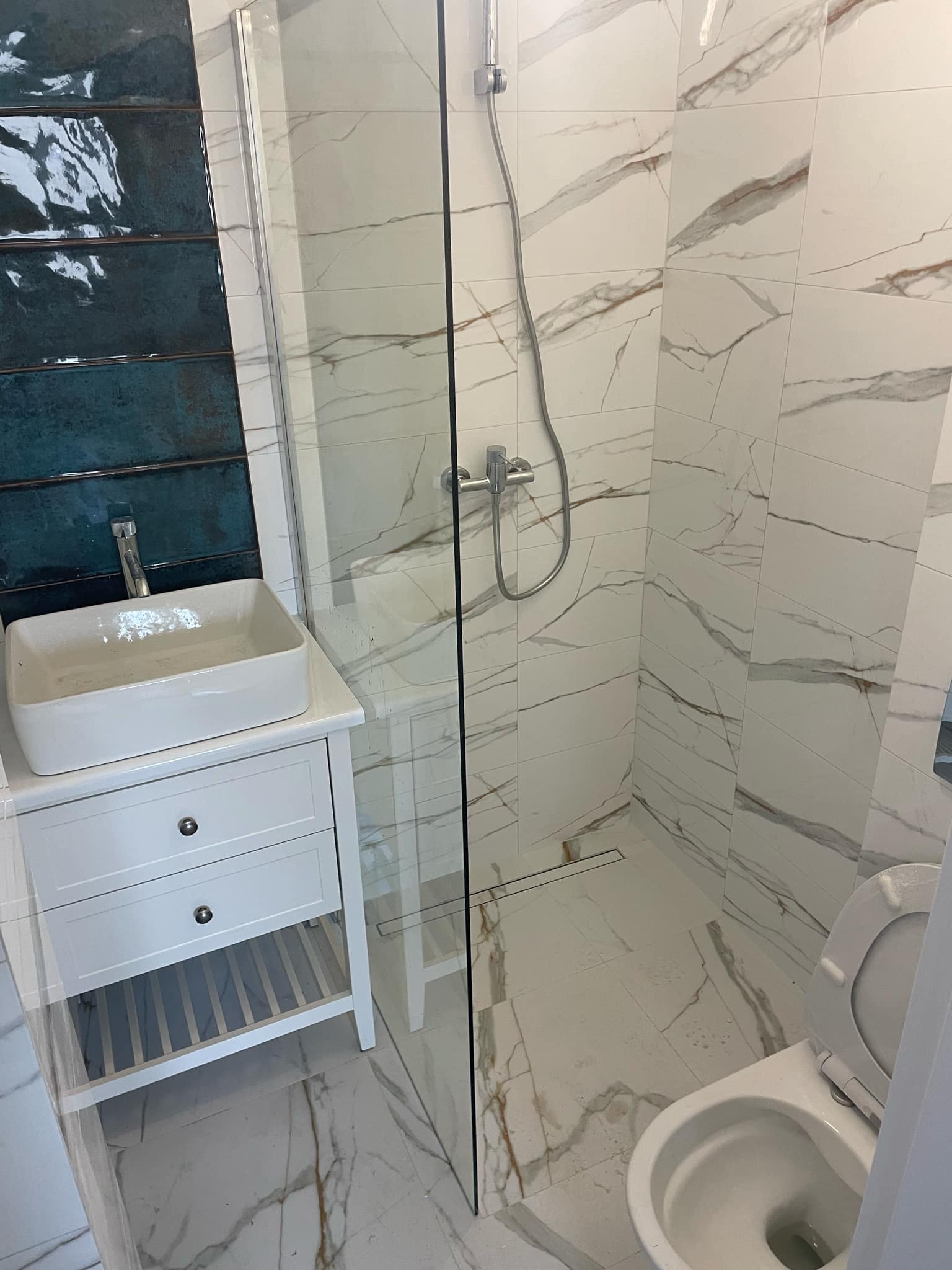 Nowoczesna łazienka po remoncie: białe marmurowe płytki na ścianach i podłodze, kabina prysznicowa ze szklaną ścianką, umywalka nablatowa na białej szafce z szufladami oraz toaleta z otwartą klapą.