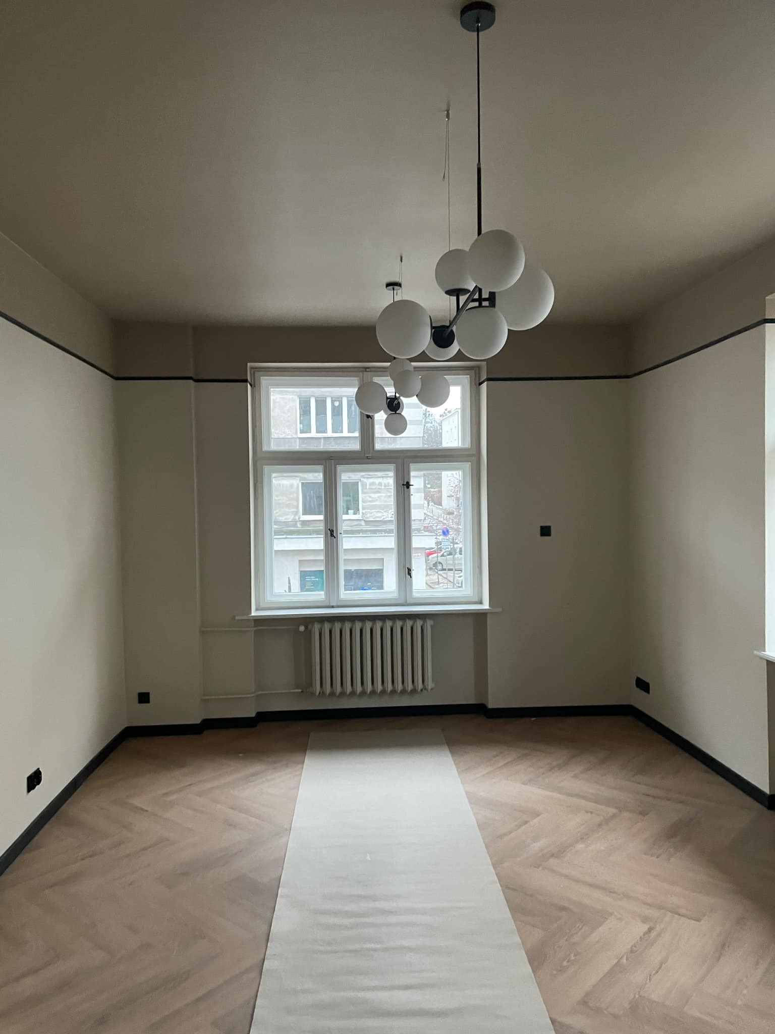 Pokój po malowaniu ścian na beżowo z ciemnymi akcentami przy suficie i podłodze, parkiet ułożony w jodełkę zabezpieczony papierem malarskim, nowoczesna lampa wisząca z białymi kulami.