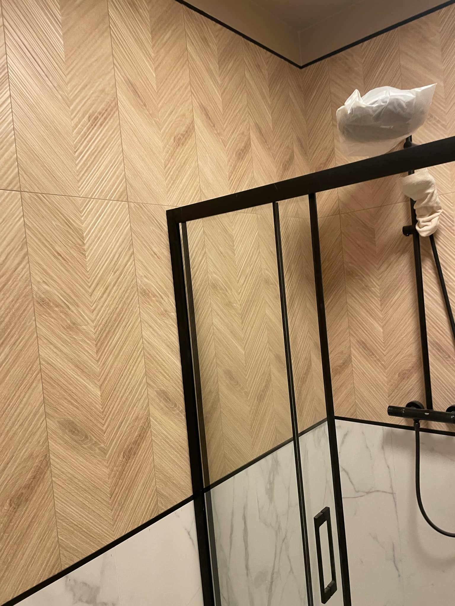Łazienka z drewnopodobnymi płytkami ułożonymi w jodełkę na ścianie, kabina prysznicowa z czarnymi profilami, armatura zabezpieczona folią ochronną.