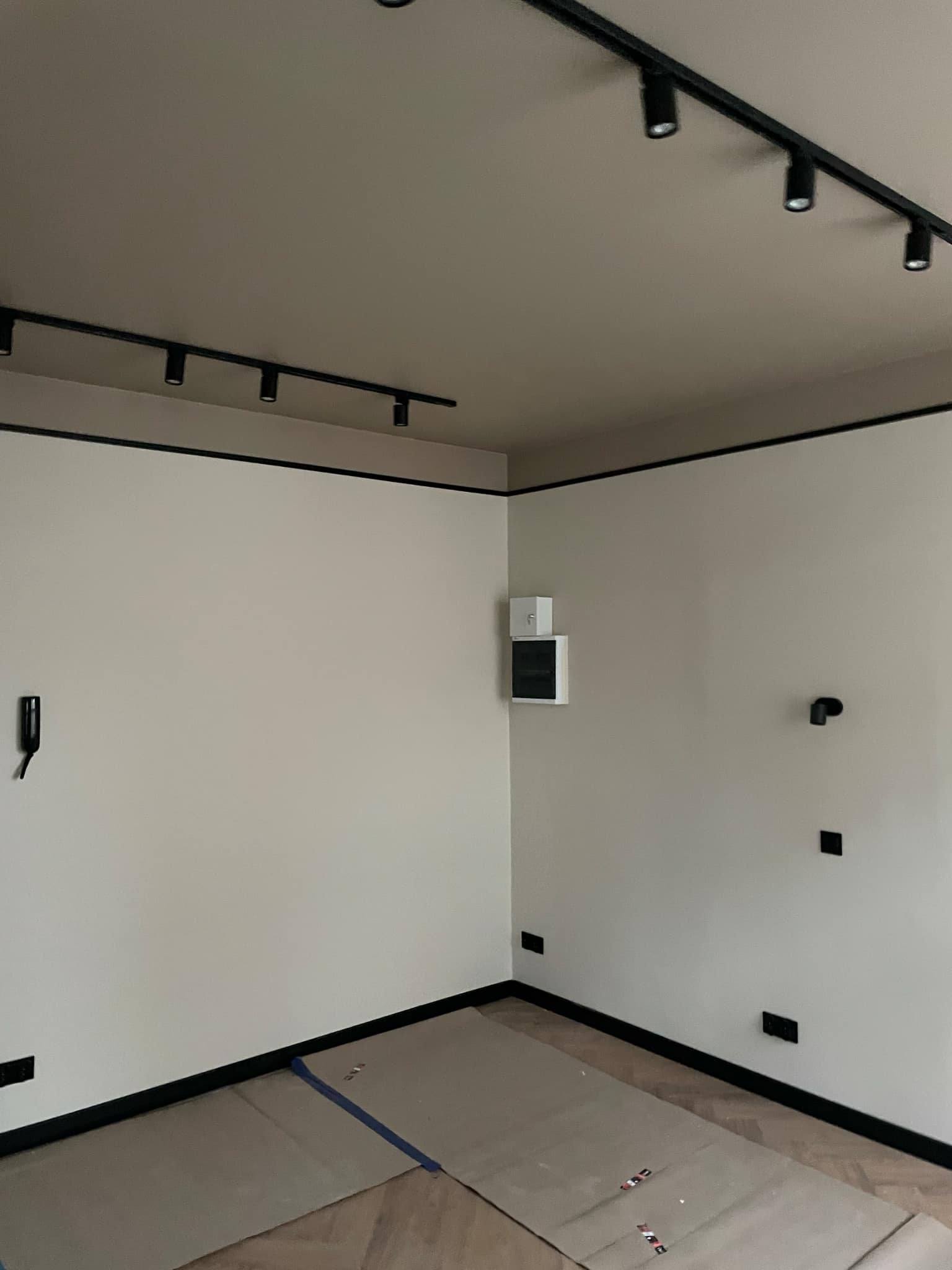 Wnętrze pokoju po malowaniu ścian na kremowo z czarnymi akcentami w postaci listew przypodłogowych i oświetlenia szynowego, podłoga zabezpieczona kartonem malarskim.