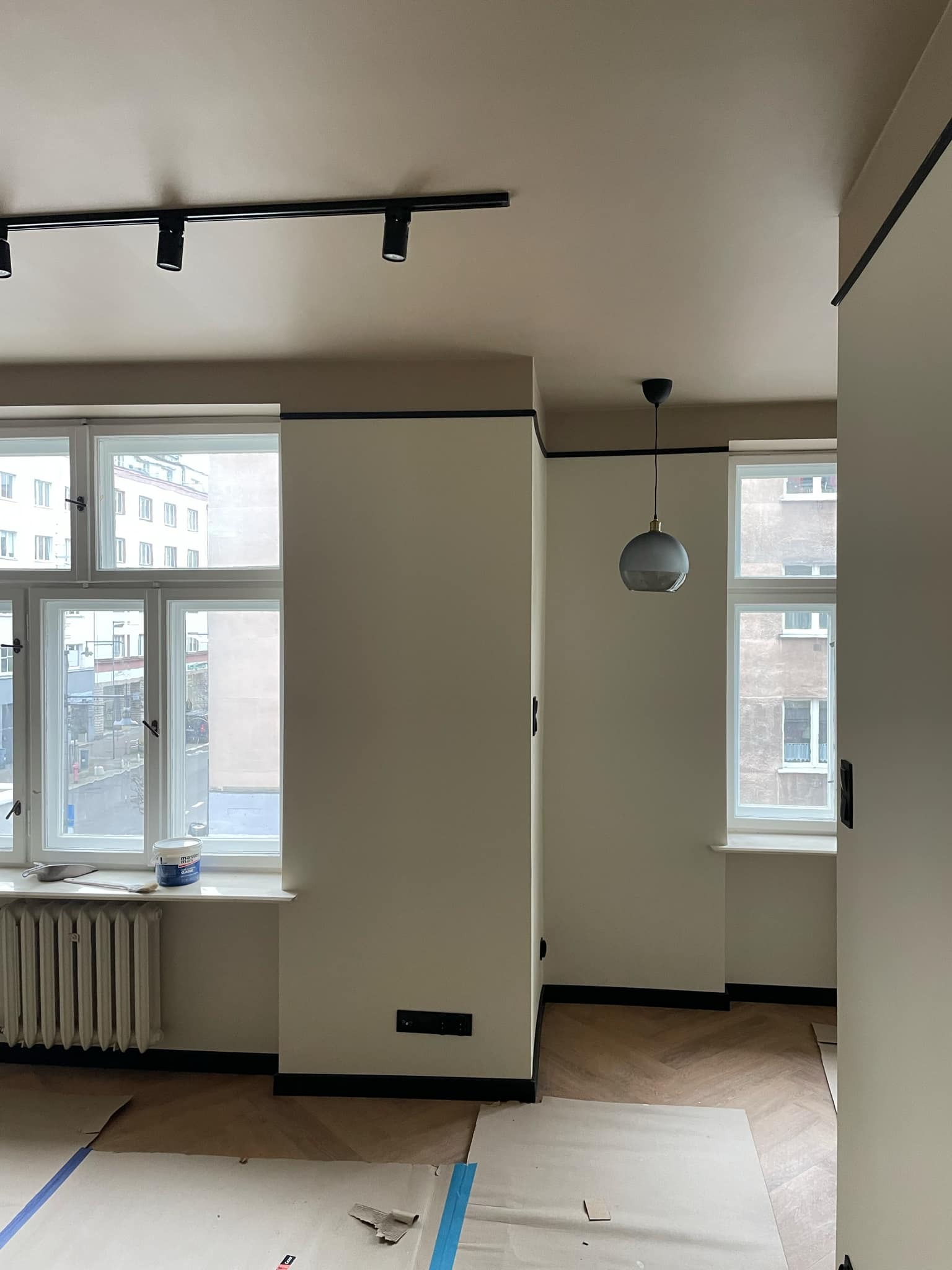 Wnętrze w trakcie malowania: beżowe ściany z czarnymi akcentami, zabezpieczona podłoga w jodełkę, puszka farby na parapecie, widok na miejski krajobraz przez okno.