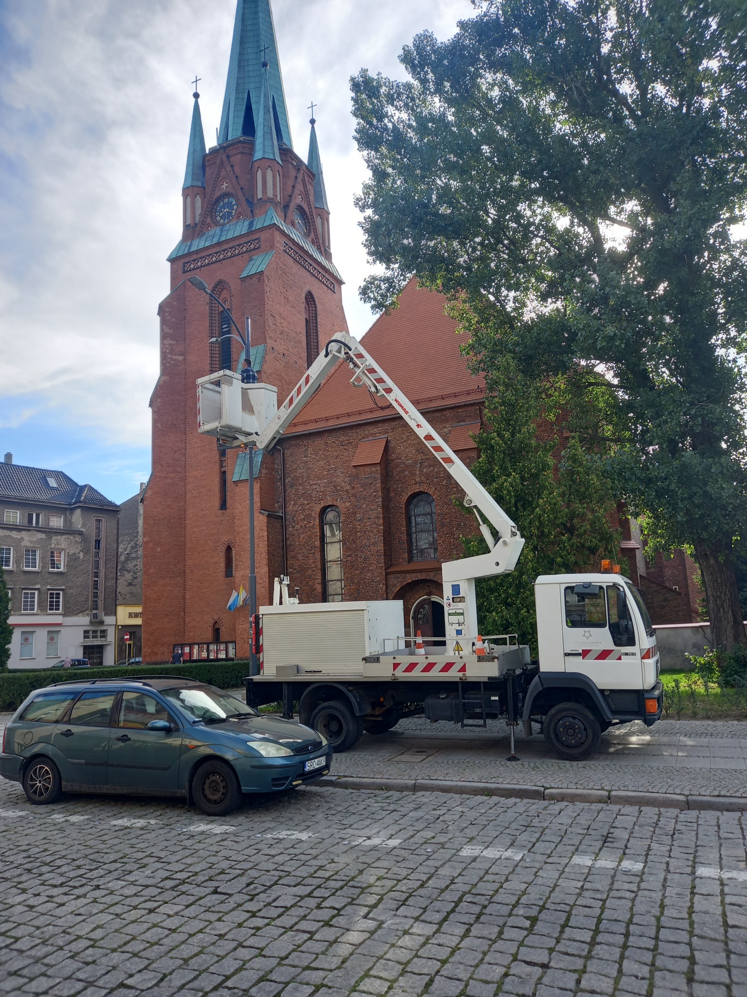 Biały podnośnik koszowy na tle ceglanego kościoła z zieloną wieżą; pracownik w koszu przy latarni ulicznej, obok zaparkowany niebieski samochód osobowy.