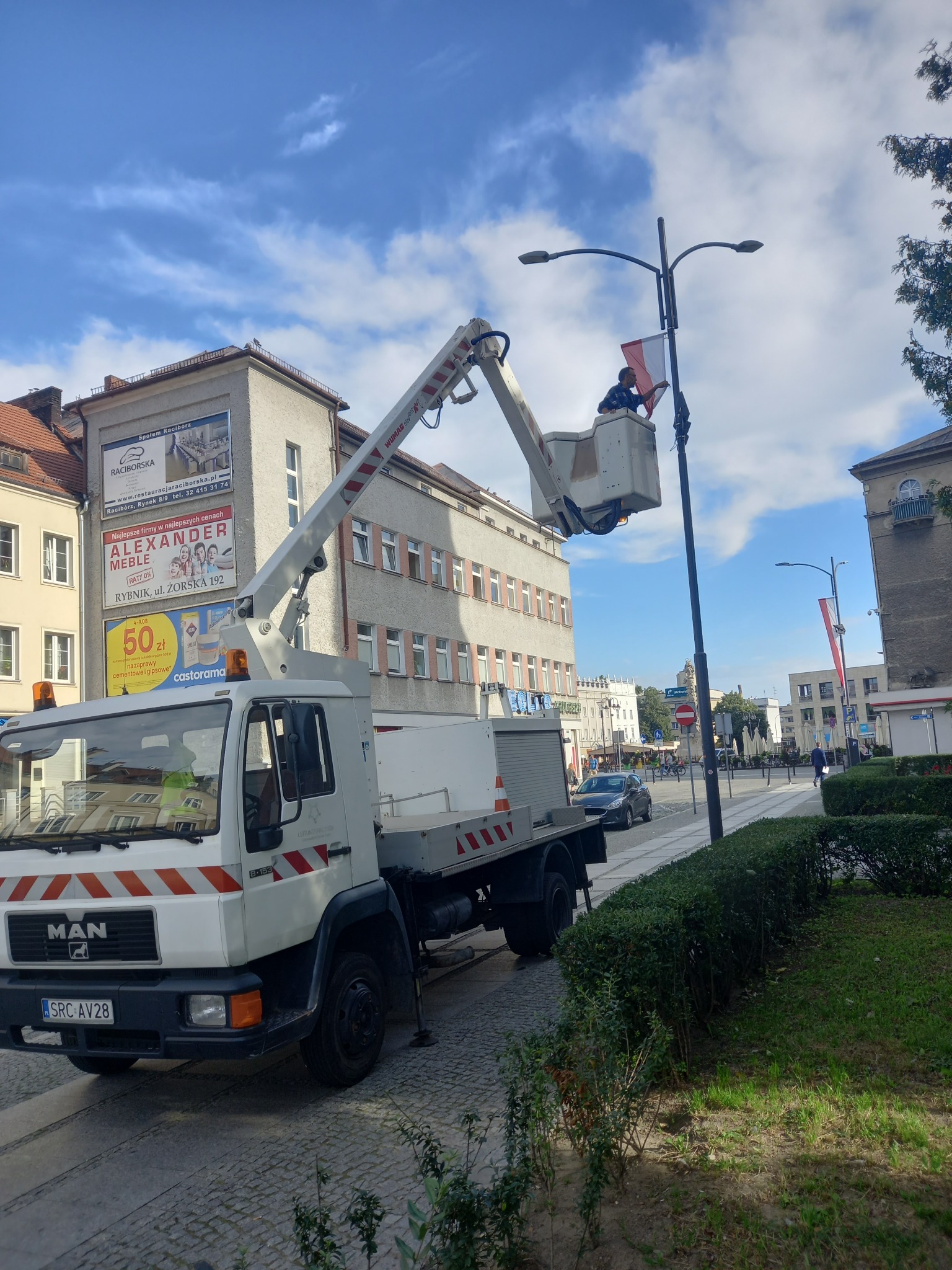 Pracownik w koszu podnośnika koszowego MAN, zawiesza flagę na słupie oświetleniowym w centrum miasta, widoczny budynek z reklamami i chodnik z zielenią.