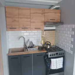 Niewielka kuchnia z drewnianymi szafkami wiszącymi, szarymi szafkami dolnymi, białymi kafelkami typu metro na ścianie, srebrnym okapem i kuchenką gazową z czajnikiem na płycie.