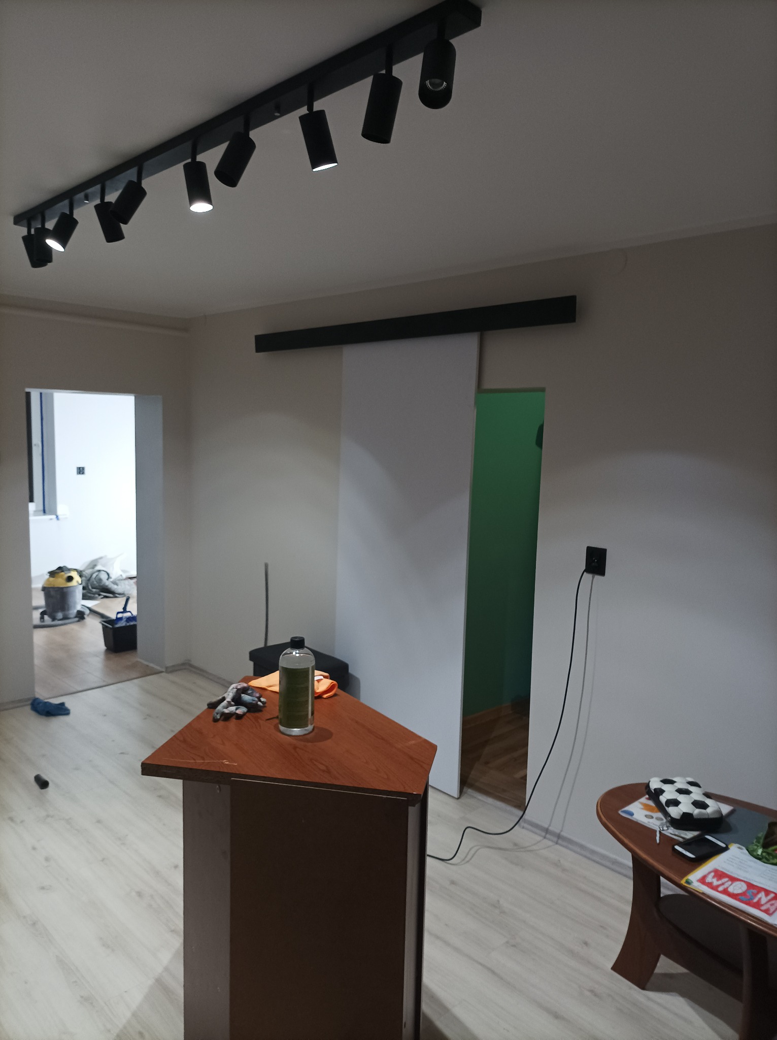 Wnętrze pokoju w trakcie prac wykończeniowych, widoczna zamontowana szyna z reflektorami na suficie, przesuwne drzwi, stolik z narzędziami i środkiem czyszczącym oraz odkurzacz przemysłowy w tle.