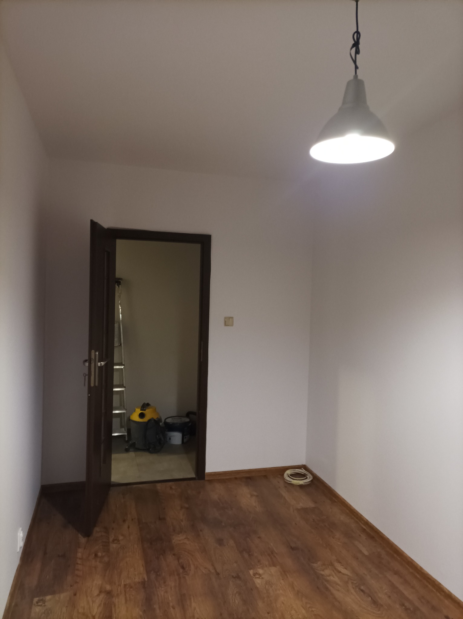 Pusty pokój z drewnianą podłogą, otwartymi drzwiami ukazującymi drabinę i odkurzacz, oraz wiszącą lampą w centralnej części sufitu. Ściany są białe, a w rogu leży zwój kabla.