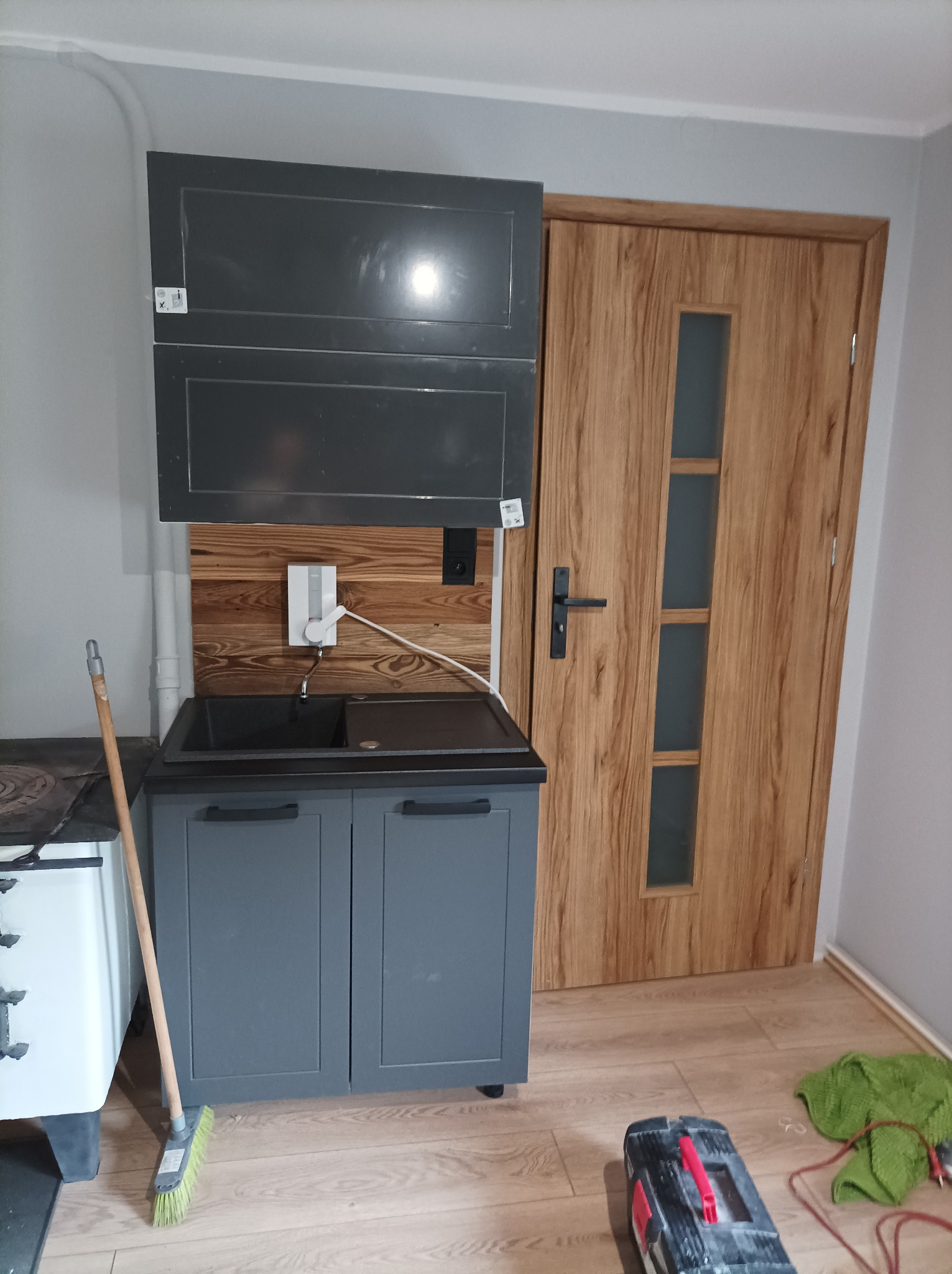 Szara szafka kuchenna z czarnym zlewem i baterią zamontowana na tle drewnianej ściany, nad nią wiszące szafki, obok drewniane drzwi z szybami, na podłodze skrzynka z narzędziami i zielona ścierka.