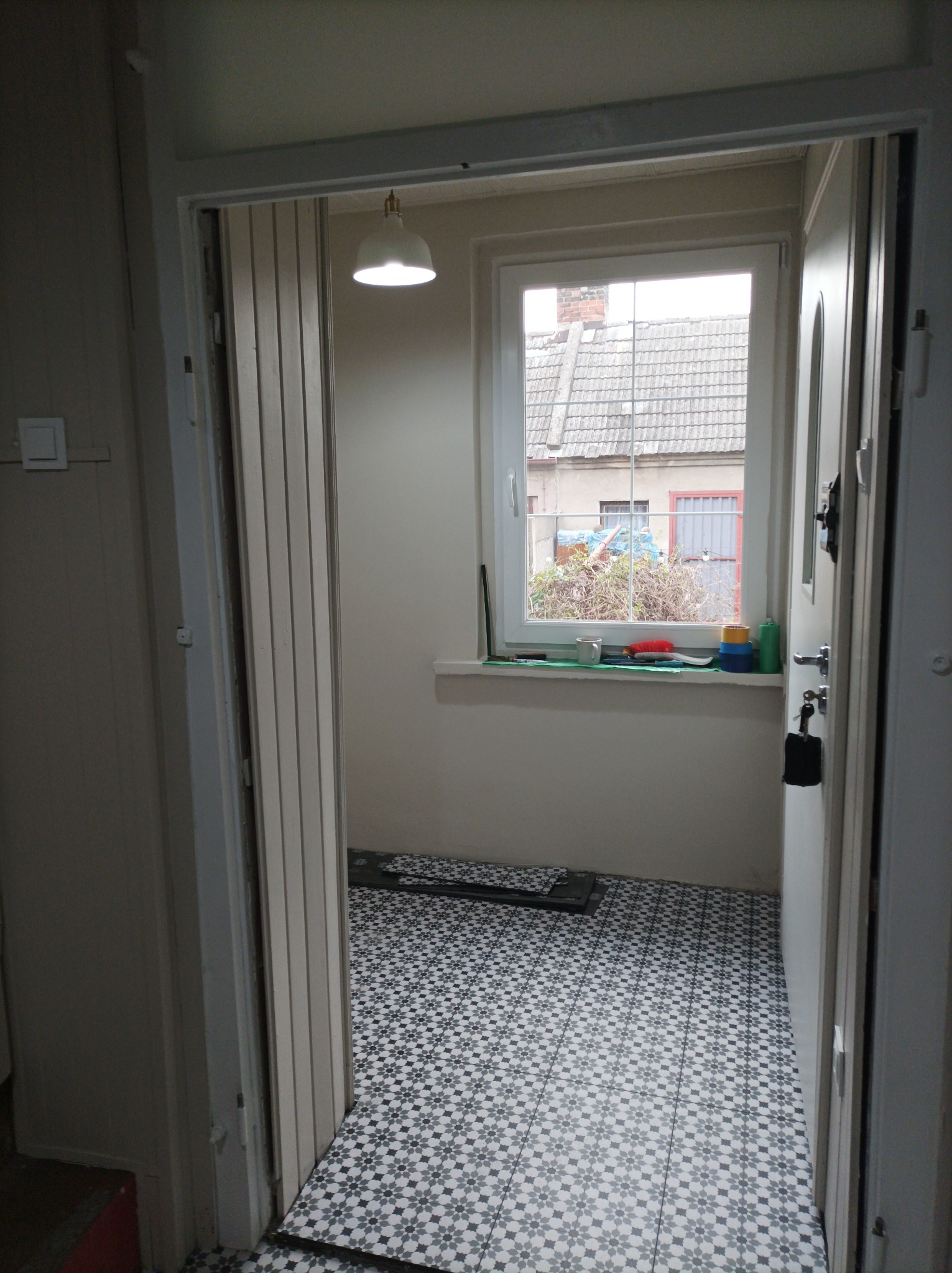 Wnętrze małego pokoju z nową, wzorzystą, czarno-białą podłogą, widok przez otwarte drzwi, na parapecie okna widoczne pojemniki i przybory.